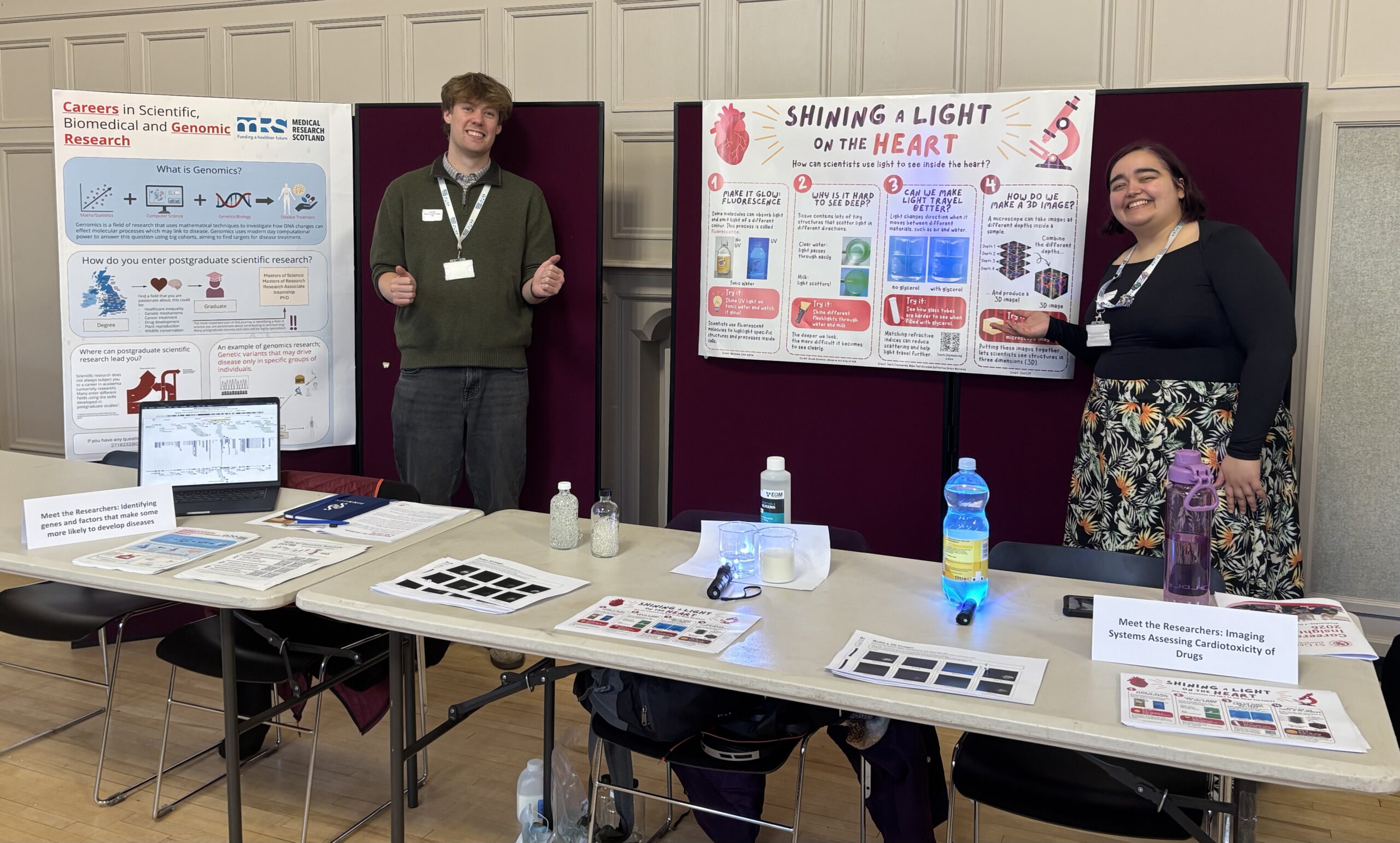 Reece and Dimona are standing behind tables on which scientific demonstrations are laid out. They are standing beside their scientific posters smiling.